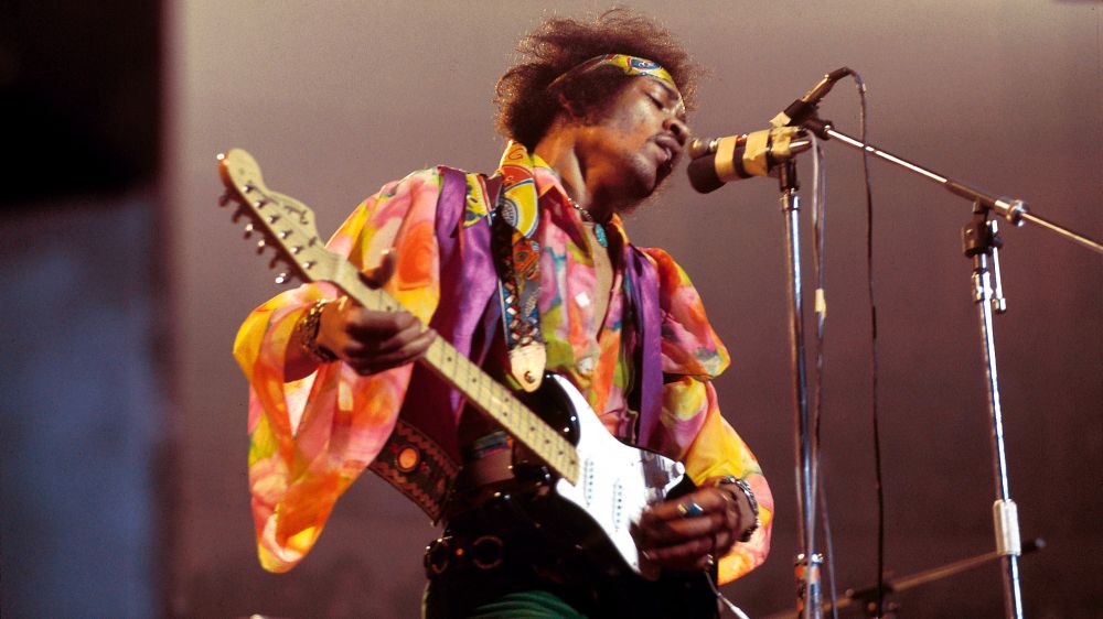 Jimi Hendrix, wearing a flowing orange blouse and playing an electric guitar.