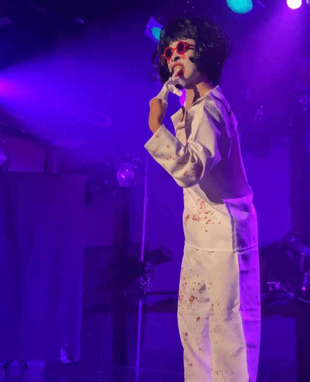 パフォーマー/コレオグラファーのいーちゃん
白塗りメイクに赤いフレームの丸いサングラス、黒髪のボブ、血飛沫が散った白いつなぎを着ている