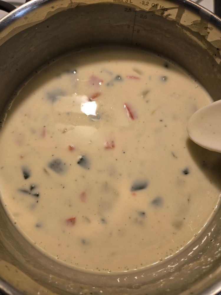 A creamy white soup with visible bits of red pepper and poblano pepper in a sauce pan. Part of an out of frame spoon is on the side, indicating someone should be stirring rather than taking pictures.