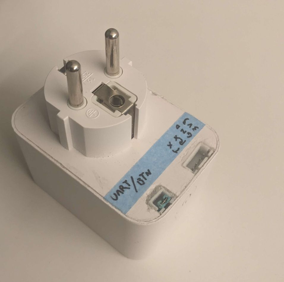 Deltaco SH-PO1E16 WLAN smart power plug, on its back on a table. the underside has been somewhat sloppily modified to have two JST-XH connectors, one 2 and one 4-pin. the 2-pin is labeled "UART/Btn" and has a jumper stuffed into it, while the 4-pin is labeled "TX, RX, GND, 3v3". the connectors appear to be superglued in place, because they are
