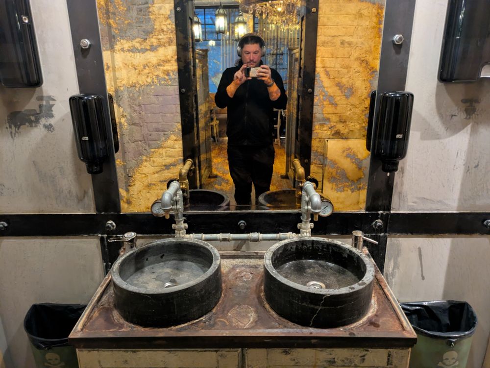 Worn out industrial style double sink. Two concrete bowls are installed on a workbench-like table. The taps are made of thick old pipes with manometers. Thick iron lathes with large bolts frame a heavily oxydised mirror and the sink bench. The wall is covered with worn out bare plasterboards