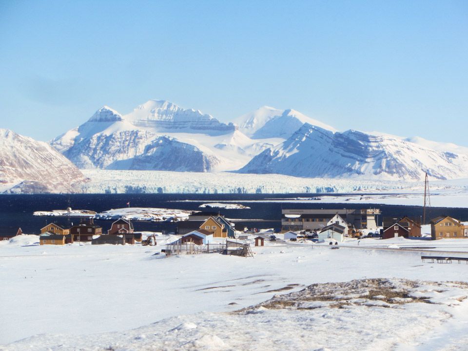 Photo of some buildings of Ny Ålesund, including NPI's Sverdrup station village with Kongsfjorden and the Loven Island, Kronebreen and mountains in the background.