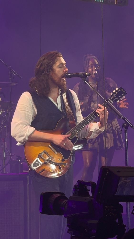 The artist Hozier performing live. He has a suit vest, long hair half-up, holding a guitar