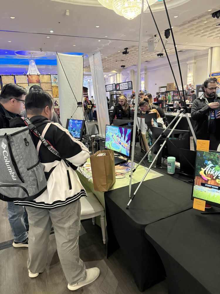 2 people playing the Capy Castaway demo at VGLX. There are black desks and screens in the forefront, with people playing games and booths in the background.