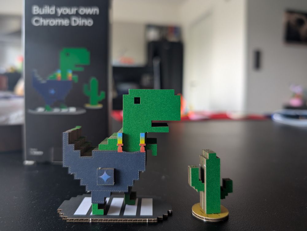 A chrome Dino with blue pants and cactus in front made out of glued cardboard 
