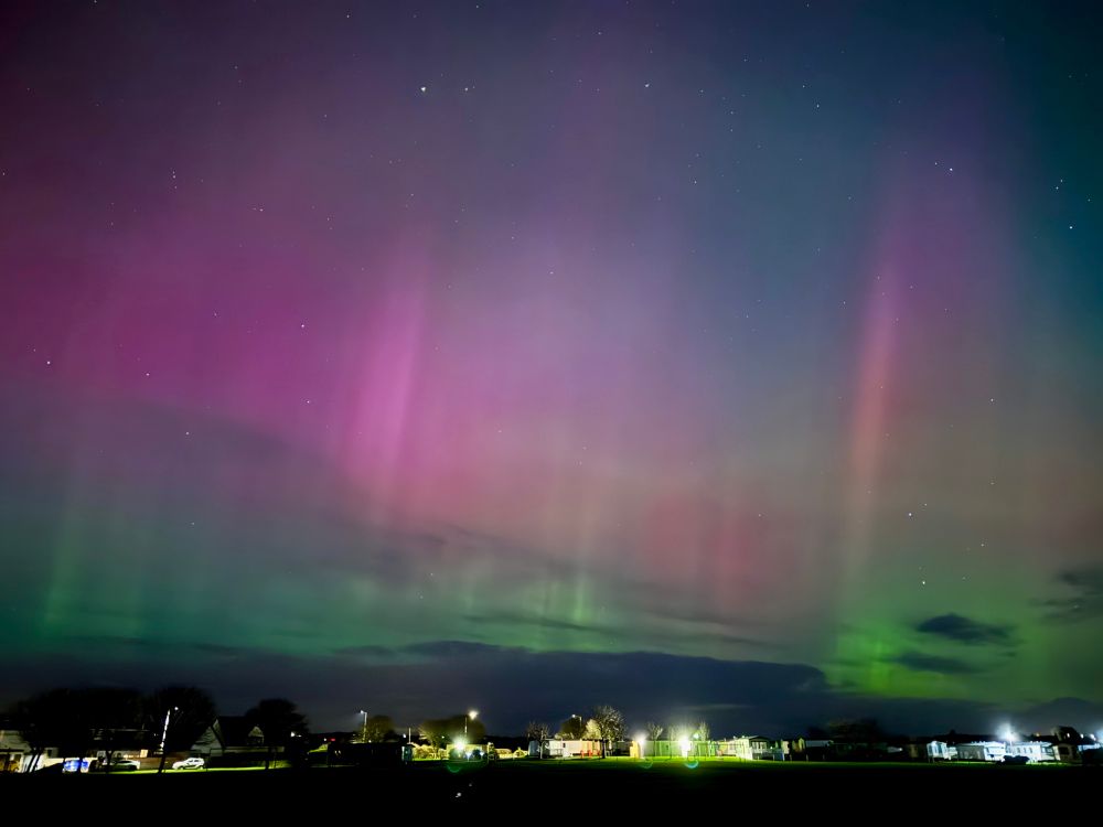 Purple and green Northern Lights in the night sky over some lit village houses.