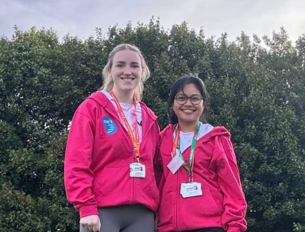 Hazelle Ongcoy and Caillee McHugh standing with green trees behind them, wearing matching pink hoodies.