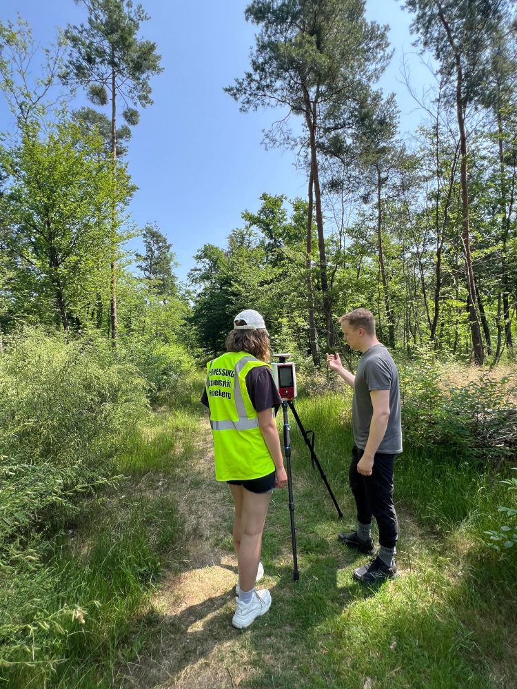 The lecturer explaining to a student how terrestrial laser scanning works. They are standing in front of the scanner mounted on a tripod in the forest.