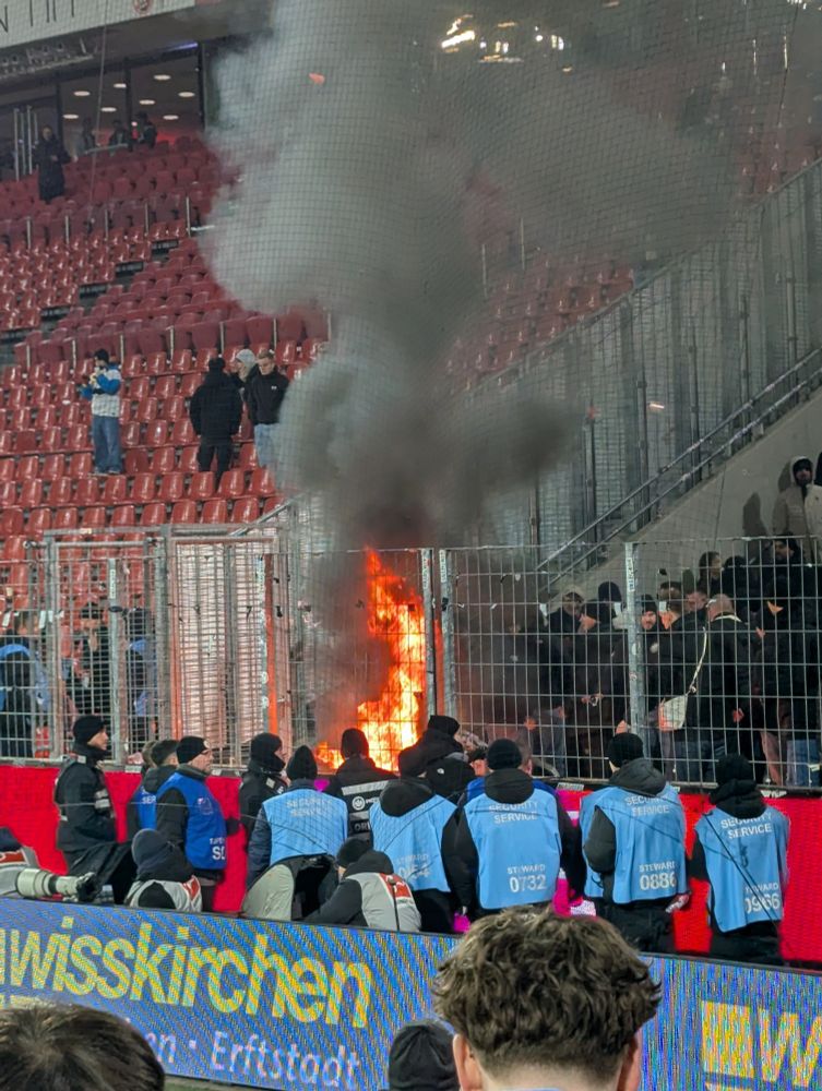 Bild vom Gästeblock im Rhein-Energie-Stadion, in welchem vorne am Zaun etwas brennt 