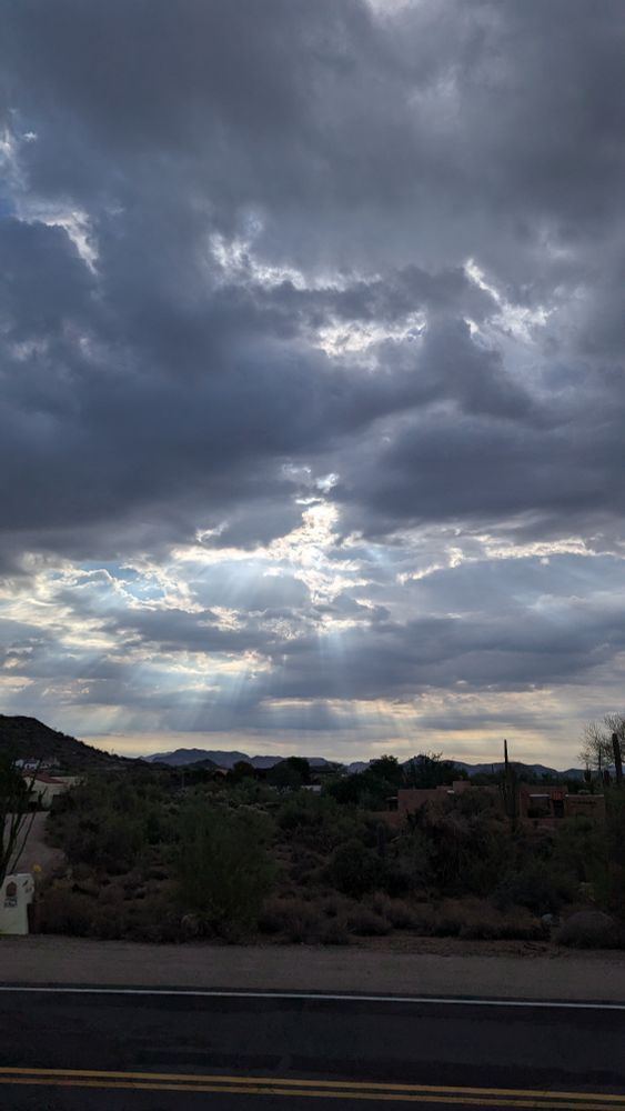 Dark storm clouds with sunlight breaking through. The button of the image is a road and shrubs, with mountains in the distance 