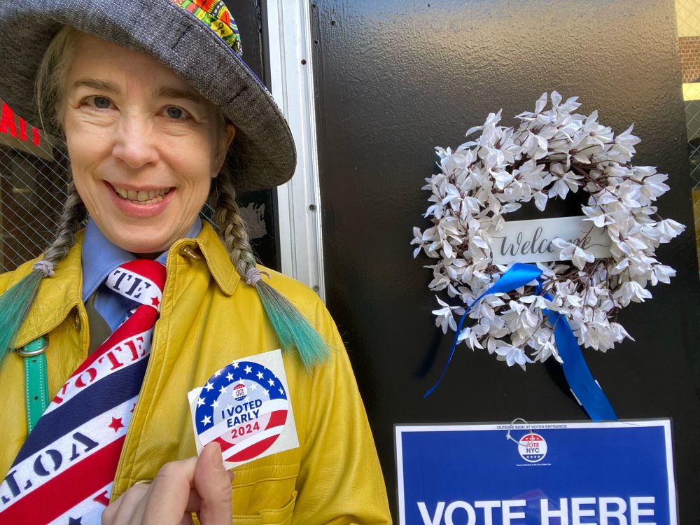 Middle-aged, grey-haired (with greeny blue tipped braids) white woman in 1960s yellow Bonnie Cashin coat, contemporary Lisa McFadden hat & 1970s red, white & blue VOTE tie in front of the door to a downtown Manhattan early polling site with a large VOTE HERE sign on the door. She's holding an I VOTED EARLY 2024 sticker. Which is a boring variation on the USA flag & was clearly not designed by a creative child. 