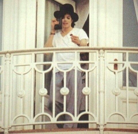 Michael Jackson stands between white double doors on a white railed balcony in a white V-neck t-shirt, a black fedora, and gray sweatpants. 