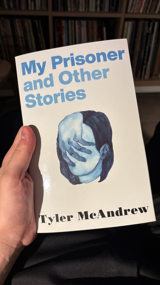 A middle-aged white guy’s hand holding the book of My Prisoner And Other Stories by Tyler McAndrew. The book’s cover has a sinister watercolor illustration of a pale blue hand engulfing a woman’s equally blue face.