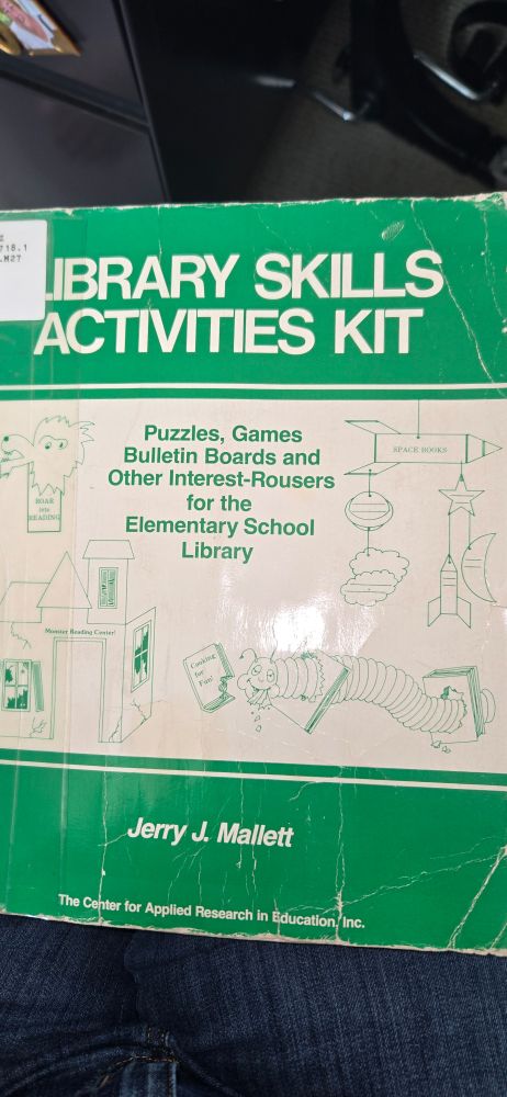 Old comb bound book titled Library Skills Activities Kits. Simple line drawings of a wonky lion bookmark, space mobile, haunted house labeled "monster reading center" and a caterpillar eating a cookbook decorate the cover. The caterpillar has half-lidded eyes pointing in different directions and a droopy antenna. Crumbs of paper fall from its smiling open mouth. At first glance it looks intoxicated, but looking longer reveals what may be a fang and subtle malice.