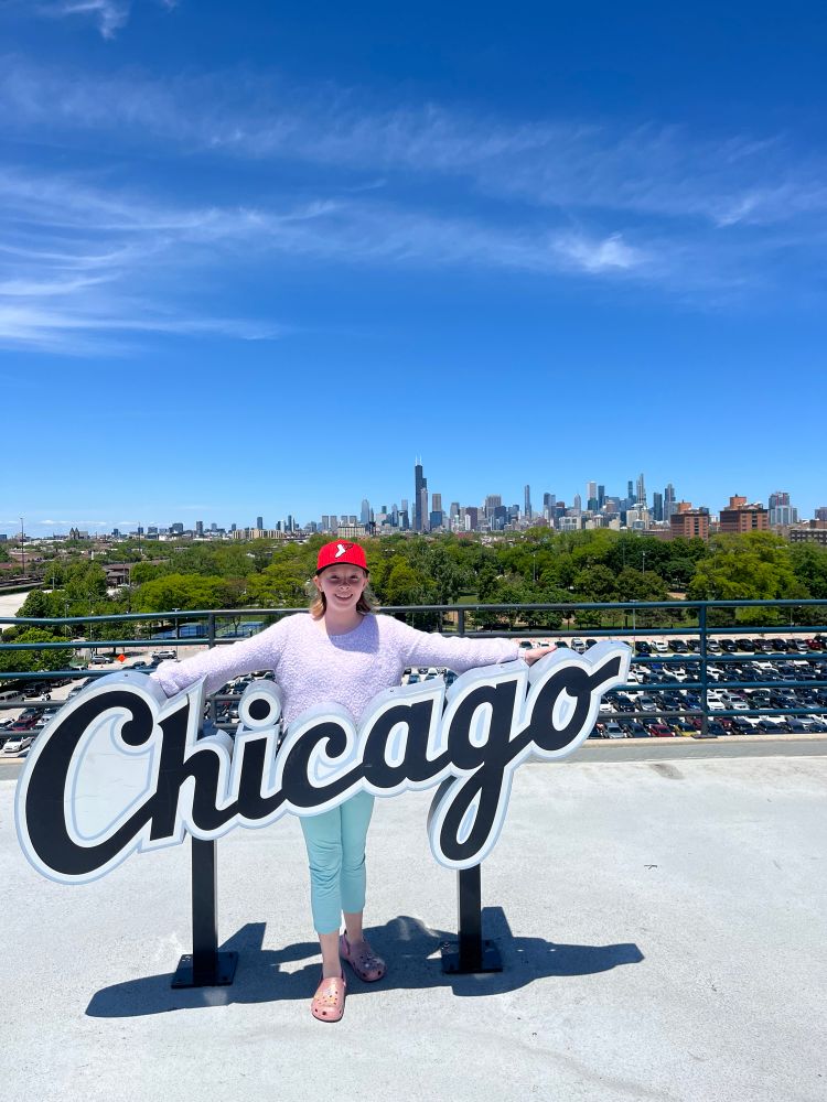Awesome kid with her arms spread out and a big smile behind a sign saying, “Chicago” in Sox-brand typeface.  Downtown Chicago is in the distance, and the sky is a wonderful blue.  