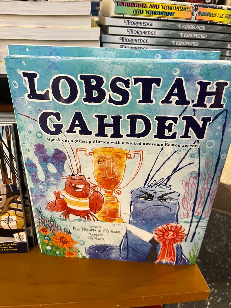 The book cover of an illustrated children’s book reads, “LOBSTAH GAHDEN”.  A red lobster standee next to an orange trophy that reads “#1”.  A blue lobster stands in the foreground with a red ribbon that reads “#2”.