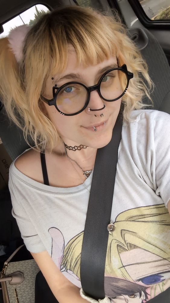 A selfie of the OP, Myrtle Faust sitting in the passenger seat of a car with their seatbelt on. Their blonde hair is in pigtails and they're heavily pierced with nostril piercings, a nose chain, septum, eyebrow and vampire bites. They have on an Ouran Highschool Host Club shirt from Hot Topic and have a black bra strap showing on their right shoulder from where the shirt falls down. They're wearing their black round kitty glasses (Judy109) from FIRMOO.com as they smile into the camera.