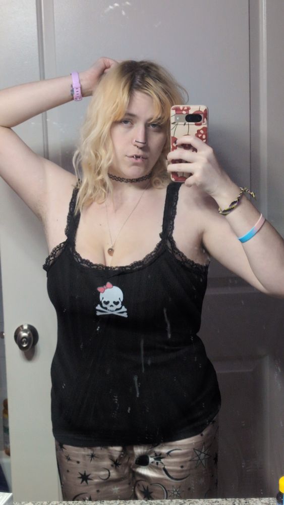 A mirror selfie of Myrtle with blonde wavy hair that ends just past their shoulders. They're wearing a black tank top with a white skull wearing a pink bow on the bust. Their pajama pants are a satin rose with black depictions of astrological symbols. Their right hands is touching the back of their head as they hold their phone (tan case, red cherries in the shape of skulls) up with their right hand to take the photo. They're looking at the camera's reflection with a doe-eyed expression. Behind them is their white bathroom door with a silver knob.