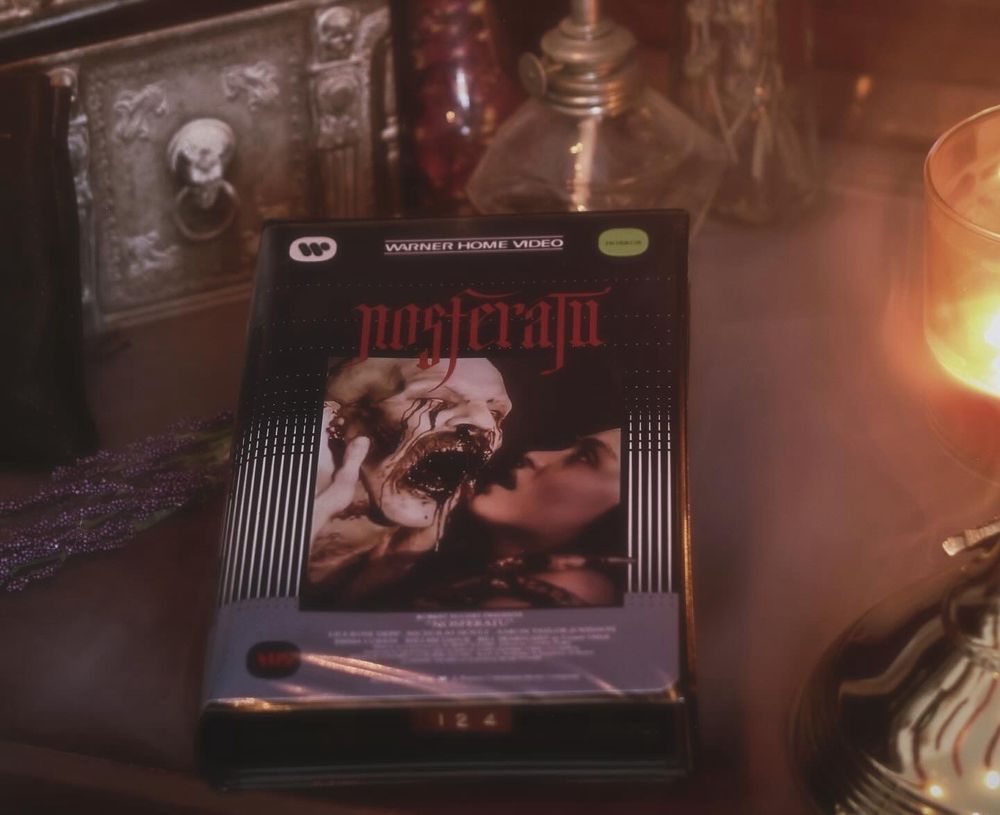 A clamshell VHS case for the Robert Eggers film Nosferatu sitting on a wooden desk surrounded by atmospheric smoke and lit dimly by candlelight 