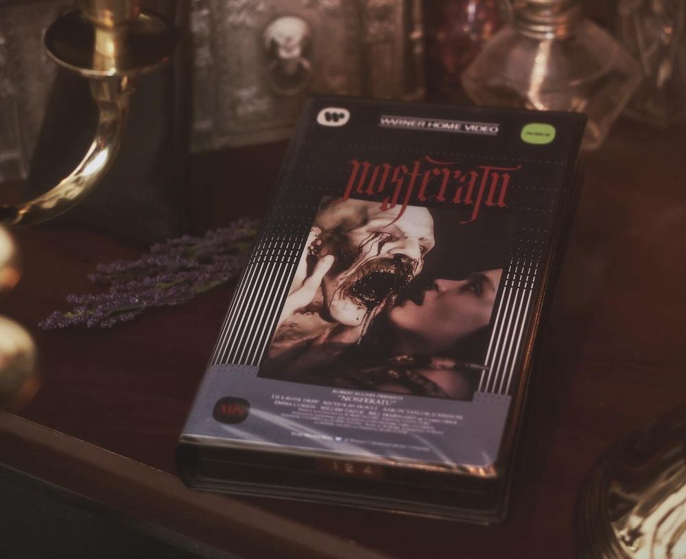 A clamshell VHS case for the Robert Eggers film Nosferatu sitting on a wooden desk surrounded by atmospheric smoke and lit dimly by candlelight