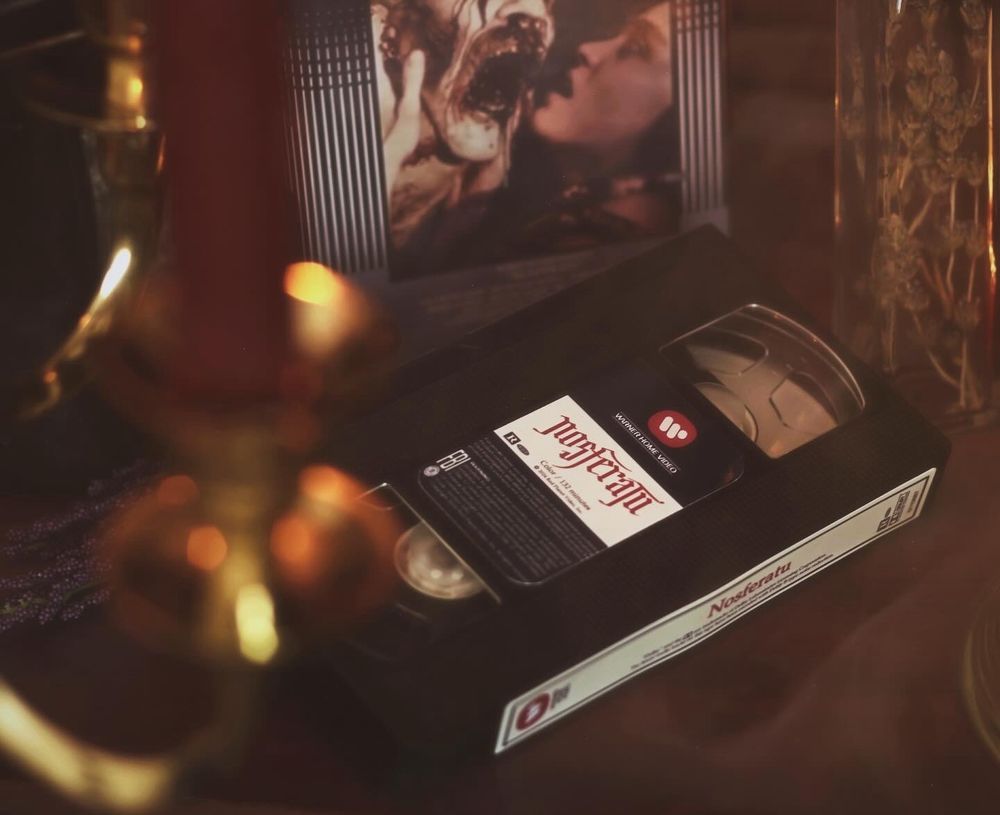 A clamshell VHS case for the Robert Eggers film Nosferatu sitting on a wooden desk surrounded by atmospheric smoke and lit dimly by candlelight