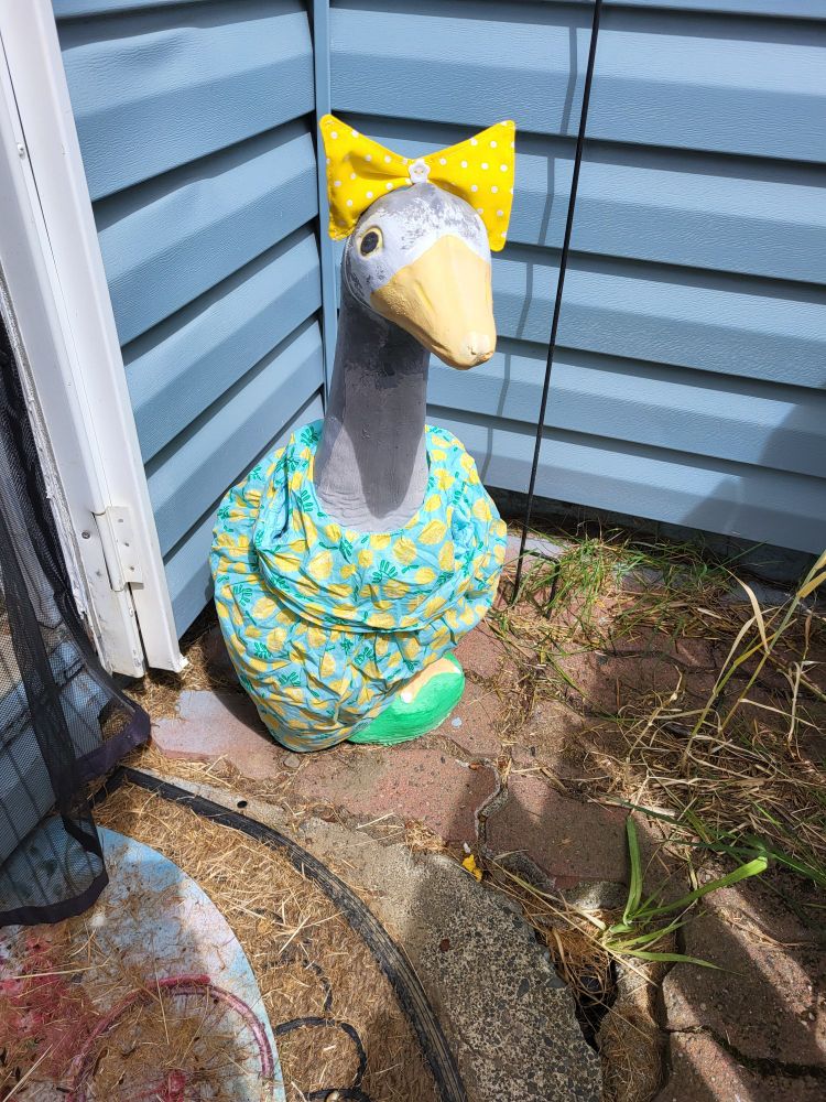 A grey concrete porch goose sits in front of a blue house. He's wearing a yellow bow with a blue sundress with pineapples on it