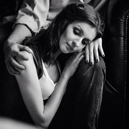 Black-and-white photograph of a woman resting her head on a man's knee. She has long, dark hair styled loosely, light skin, her eyes are half-closed, and is smiling peacefully. The man is sitting on a large chair with his arm is gently draped over her shoulder, emphasizing closeness and comfort. 

