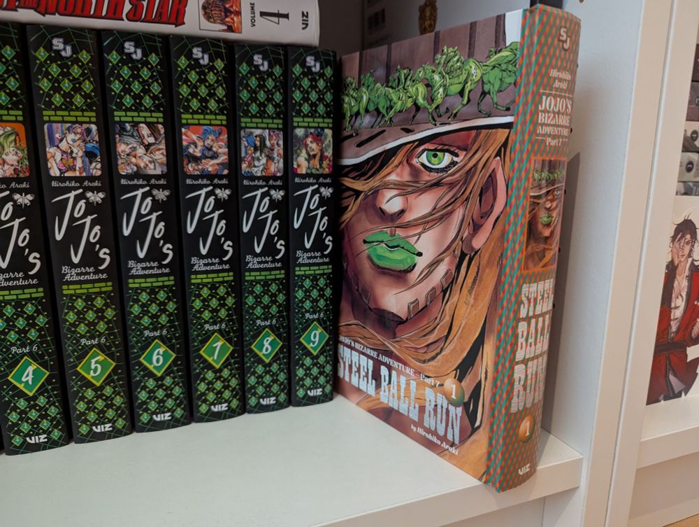 Picture of Steel Ball Run volume 1 being put in a bookcase besides other Jojo volumes.