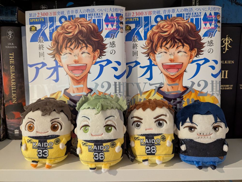 A picture of two of the same issue 30-2025 of Big Comic Spirits being displayed in a bookshelf. On the cover is Ashito, a young man with messy brown hair smiling a big open mouth smile, eyes closed. Standing in front of the magazines are small chibi like plushies of several Ao Ashi characters wearing their kit, which is yellow with a black stripe down the middle. 