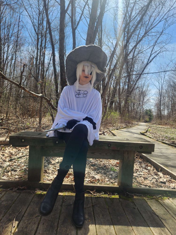 Photo of the poster cosplaying Siffrin from In Stars and Time, sitting on a bench by a tree, eyes closed