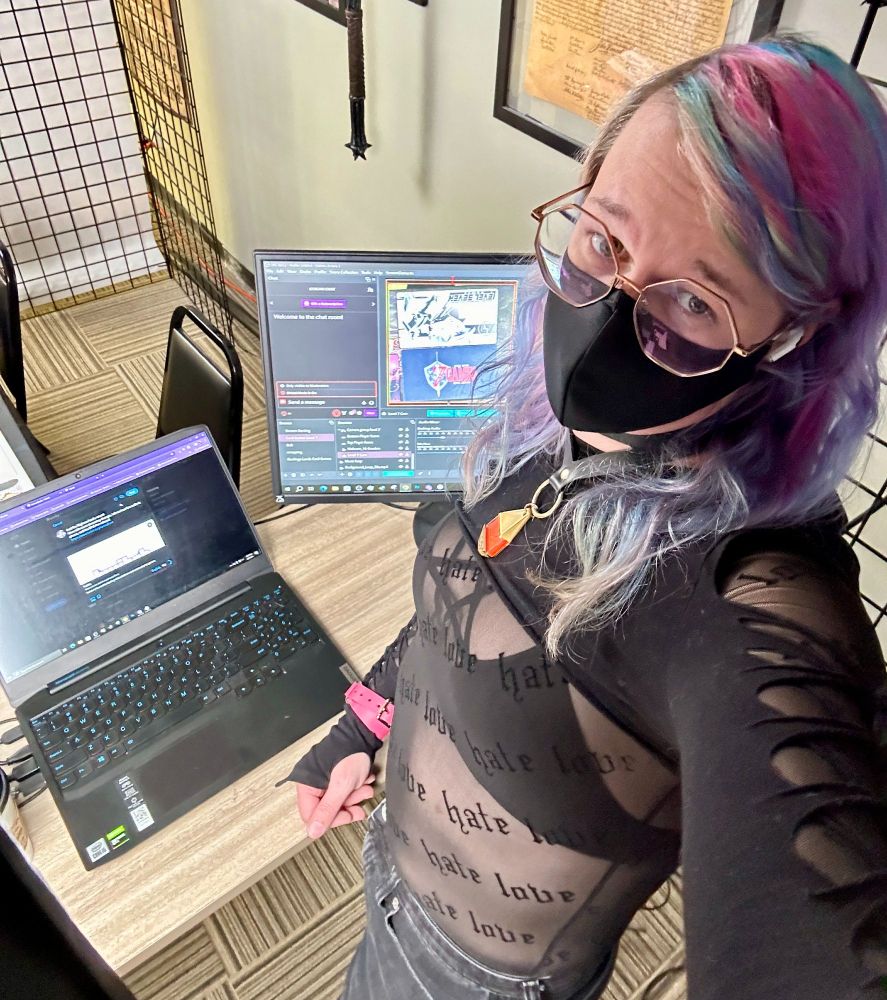 A selfie of me at a standing desk with a laptop and a second monitor displaying OBS. I'm wearing black denim shorts, a thin black mesh top that has "love" and "hate" printed into it, wearing a top under it with a pentagram, an arm laced shrug, a collar with a digimon tag, and a black mask