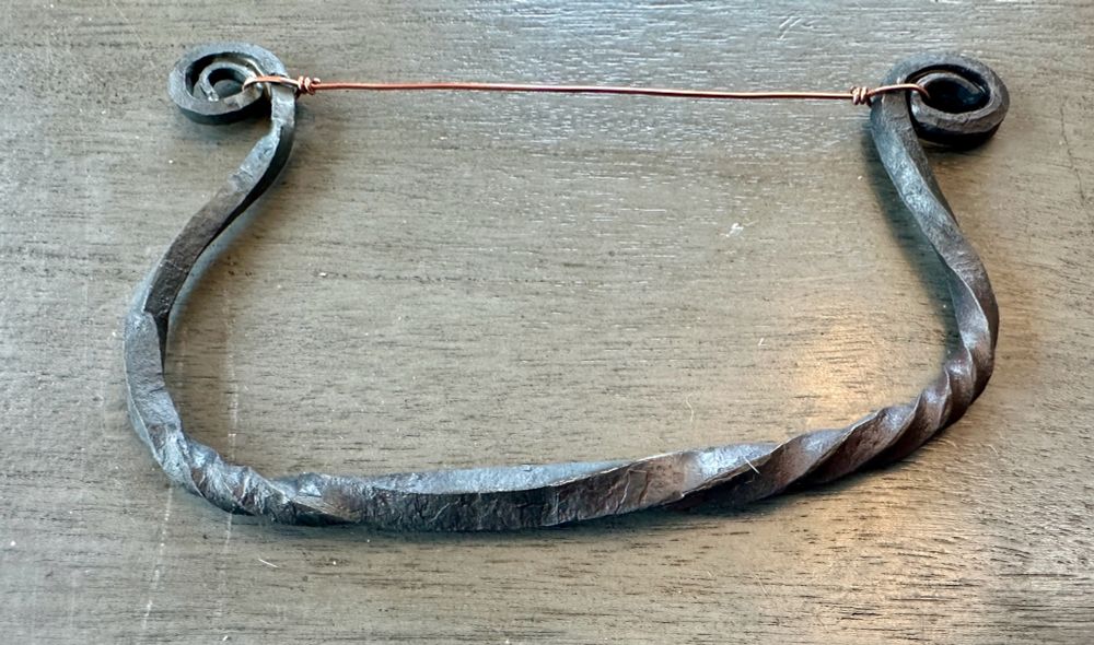 A close shot of a cheese knife made from rough black steel. It’s curved like a bow with loops and both ends and taut copper wire running between both loops.