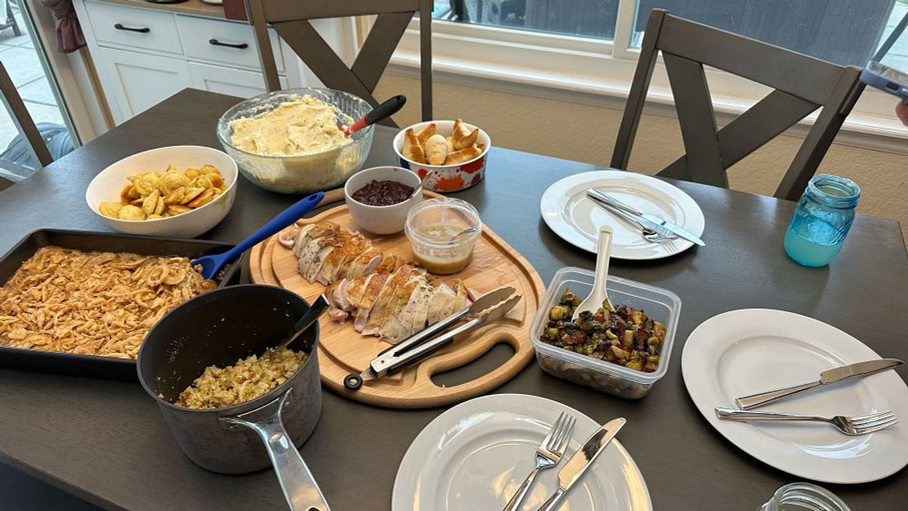 A full thanksgiving day spread, three empty plates, a wooden cutting board with turkey breast, a pot of stuffing, a Tupperware of bacon Brussel sprouts, a green bean casserole, a bowl of duchess potatoes, another bowl of mashed potatoes, and a small container of crescent breads.