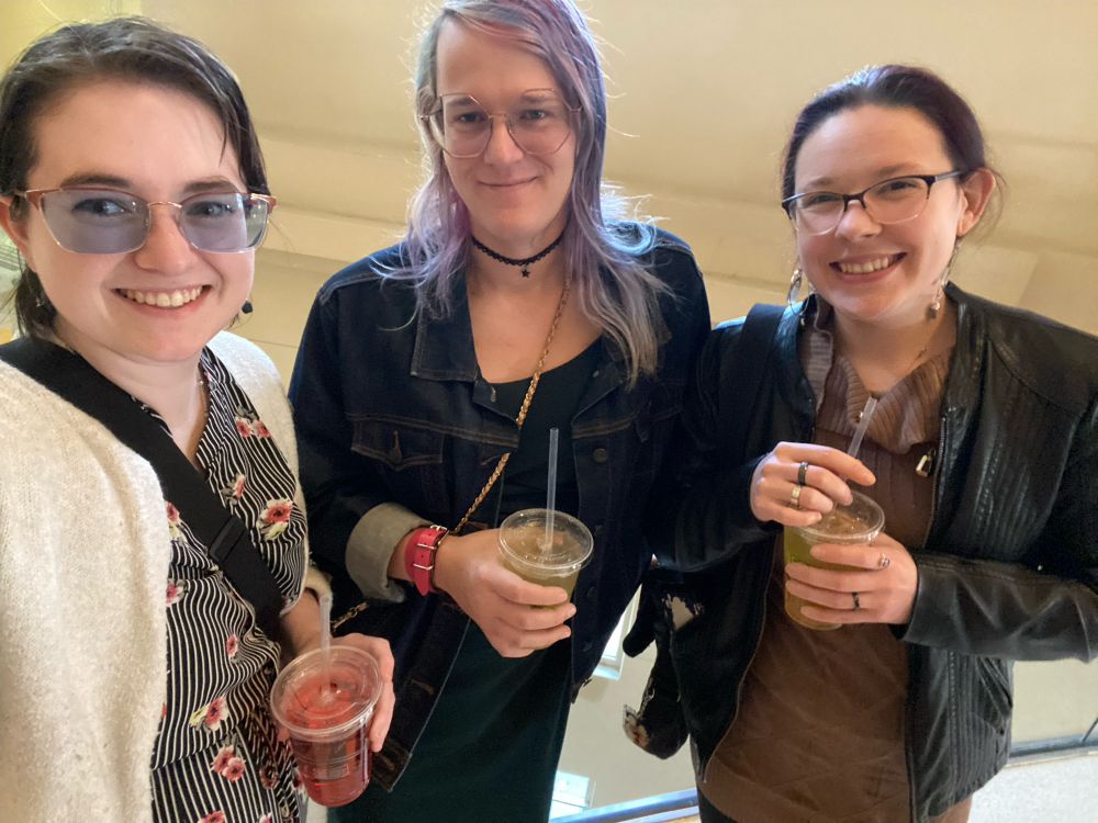 A selfie of, in left to right order, Hailey in a black and white vertical striped dress with floral accents and a white cardigan, then me in a forest green dress, denim jacket, and black lace choker, and Steph in a tight brown cowl dress and leather jacket. We’re all holding drinks and smiling at the camera.