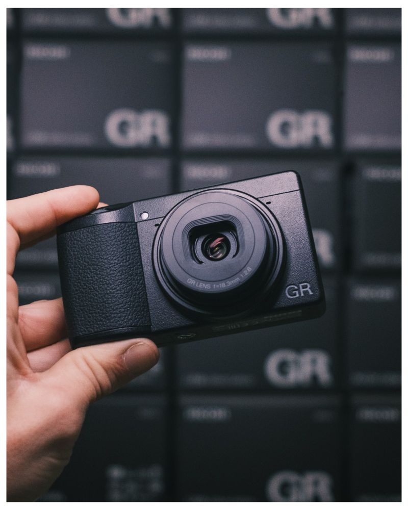 Photo of a ricoh GR III in front of a large stack of Ricoh GR III boxes. 