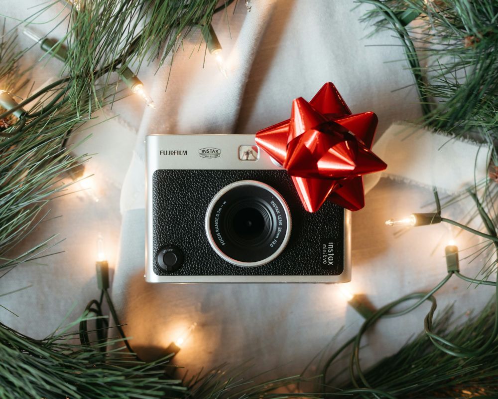 An image of a Fujifilm Instax mini Evo Camera with a red bow on it surrounded by pine needles and holiday string lights. 