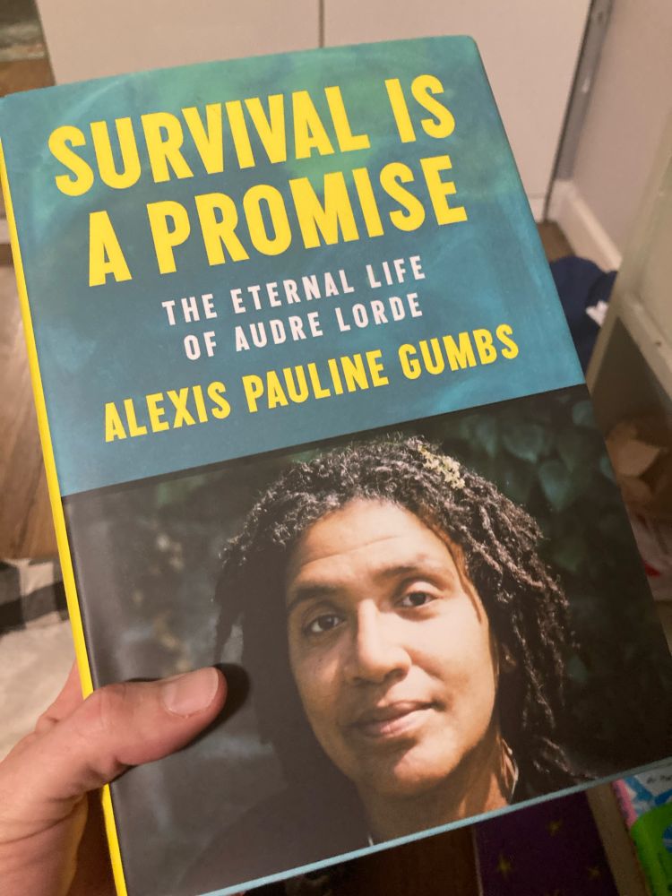 Leigh holds a copy of Survival is A Promise The Eternal Life of Audre Lorde by Alexis Pauline Gumbs with a photo of Audre Lorde on the front 