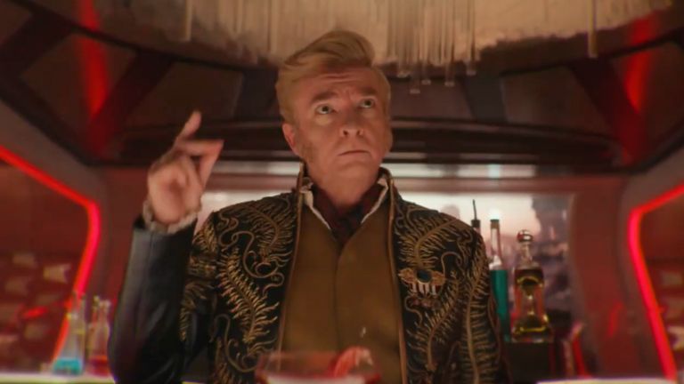 Rhys Darby as Trelane from Strange New Worlds wearing blue and gold and a sharp collar, surrounded by cocktails, snapping his fingers.