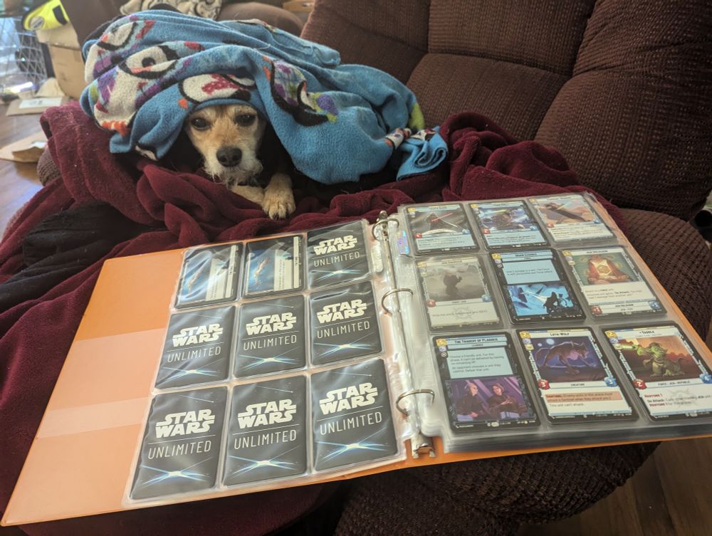 A burgundy recline with a pile of blankets on it.  A small dog is burrowed under some of the blankets, and an orange binder with many Star Wars Unlimited cards in it is on the arm of the chair.