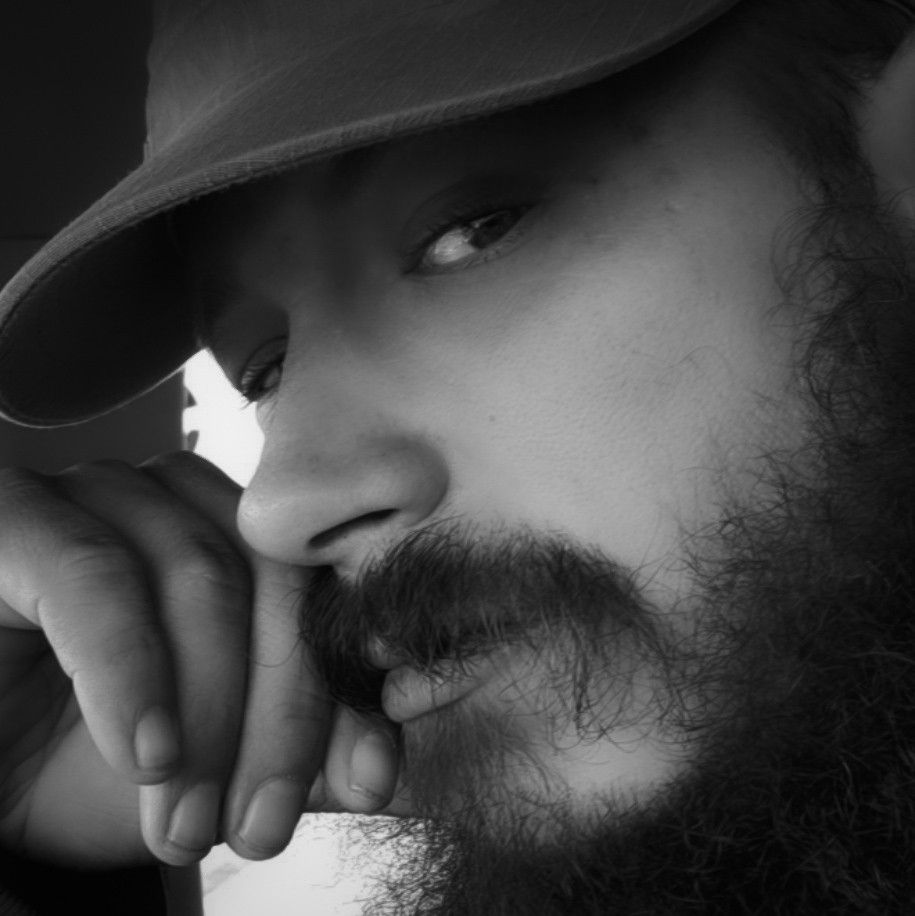 black and white close-up photograph of a masc person with a contemplative expression. Has a full beard and mustache and is wearing a cap that casts a shadow over part of face. Head is slightly tilted, and rests face on hand, fingers curled near lips