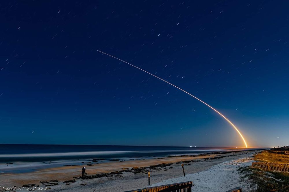 a night time beach scene. there is a bright streak of light curving up and away, the light from a Falcon rocket launch