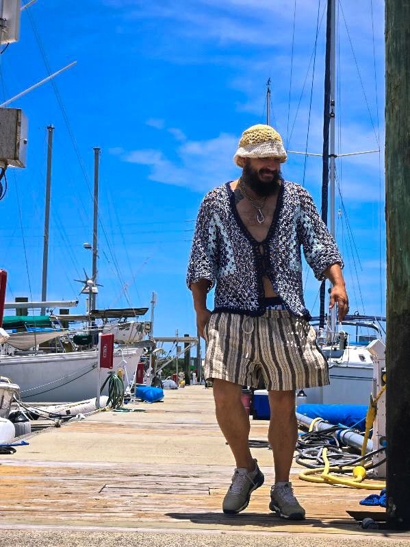 a person walks confidently along a wooden pier under a bright blue sky. They are dressed in a blue patterned open shirt, loose striped shorts, and gray athletic shoes. A beige crochet bucket hat, and they wear several necklaces. sailboats and nautical equipment in the background.