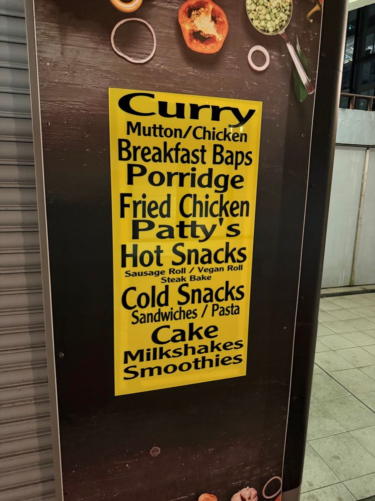 A sign on the side of a train station kiosk lists food they sell like Curry, Breakfast Baps, Fried Chicken, Patty’s, Cake and Pasta. It’s all in the same font but each entry is a wildly different size, with some of the text being squished while other words are stretched out in a comical fashion