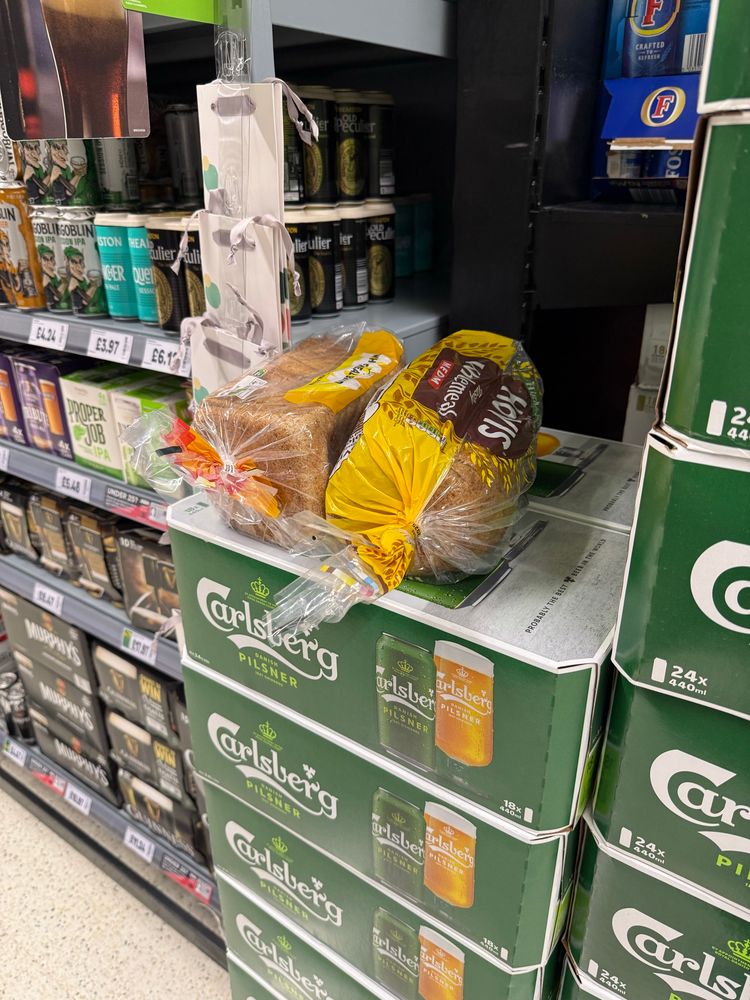 Two prepackaged loaves of sliced wholemeal bread (one is Asda’s discount brand, one is Hovis brand) have been abandoned on top of the 24-packs of cans of Carlsberg