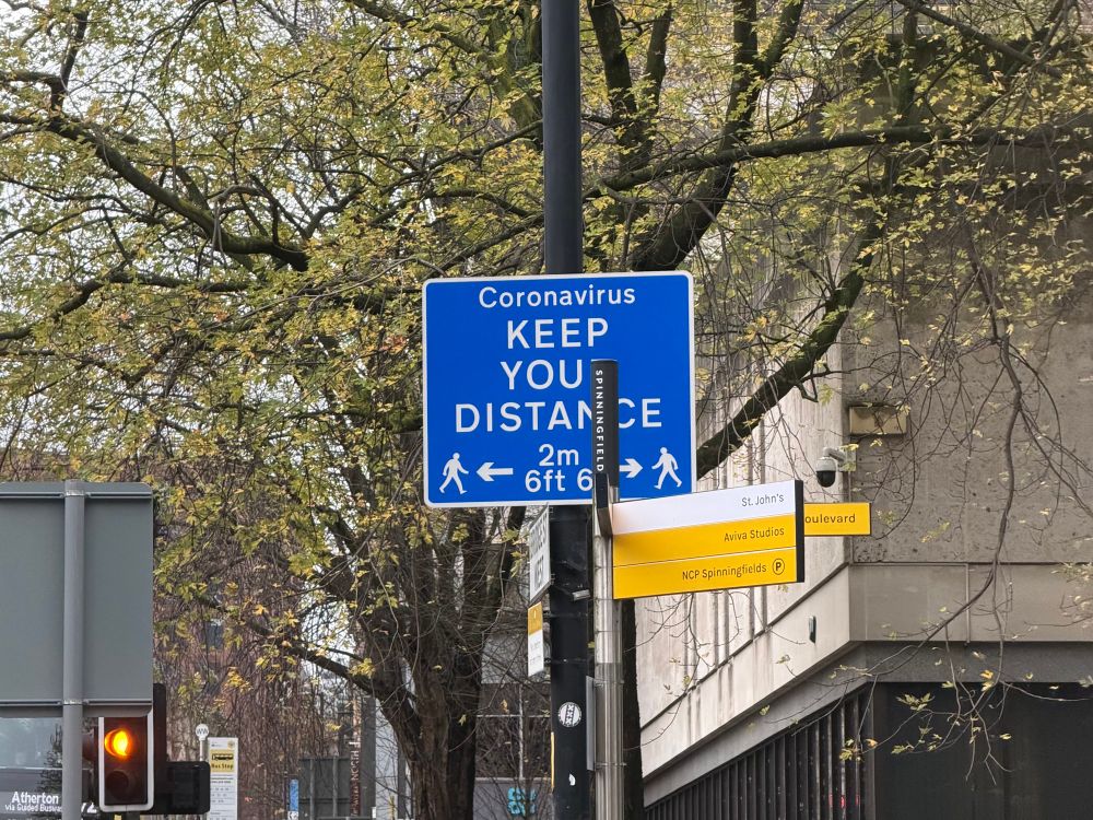 A blue sign (in the standard UK road sign style) mounted high up on a post says “Coronavirus — Keep your distance”, with a symbolic illustration of two pedestrians two metres apart