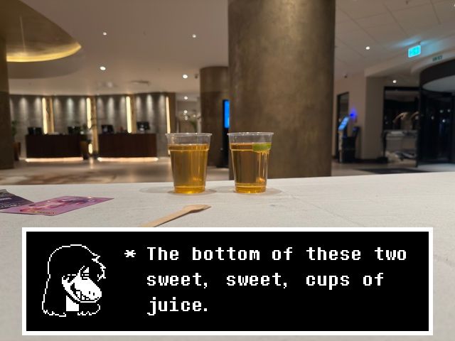 A photo of two plastic cups on a table in a hotel lobby, both filled with a very yellow liquid. One has a piece of lime in it. Overlaid is Deltarune dialogue where Susie is saying “The bottom of these two sweet, sweet, cups of juice."