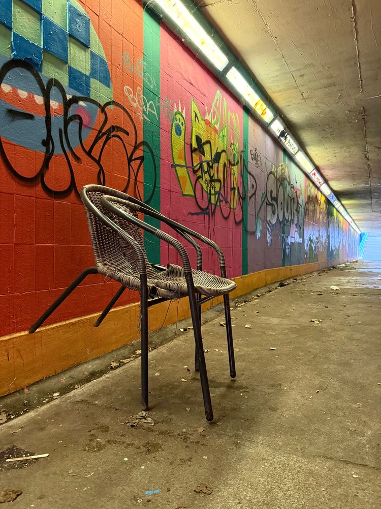Two rather damaged wicker chairs are performing some kind of mating ritual in a pedestrian underpass — one is nested inside the other, which has its rear legs balanced on the rather colourful concrete wall