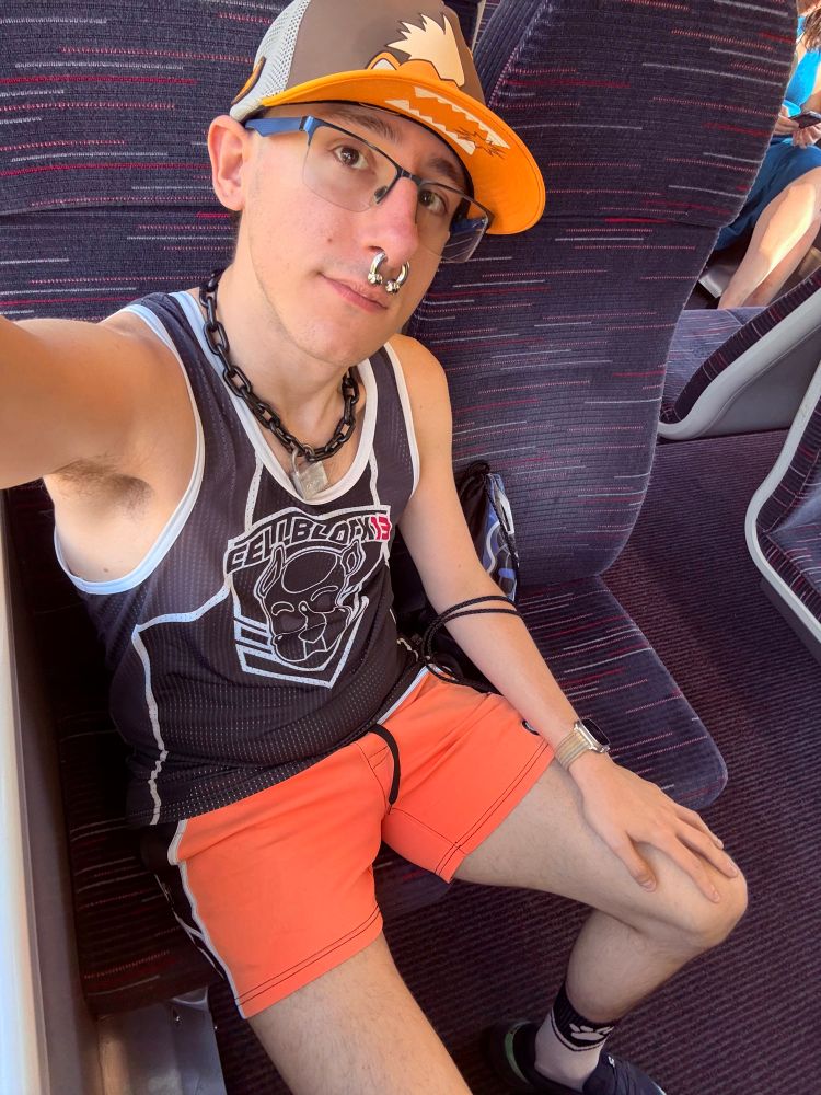 extremely gay selfie where I’m sitting on a train, angled down to show most of me — I’m wearing the Growlithe cap, a black CB13 puppy tank top, tight orange shorts, my black chain collar with a padlock, and white socks with a pawprint symbol on them