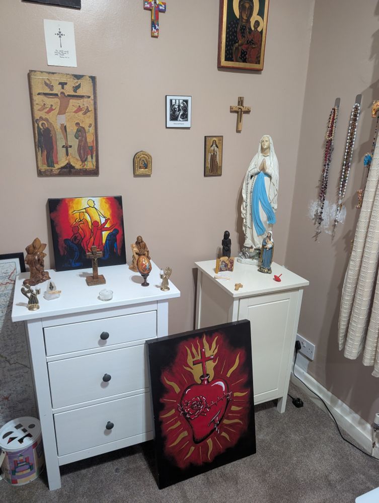 This is a prayer space. It's the corner of a room with two small white cabinets. There are religious icons on the wall. There's a statue of Mary, Mother of Jesus, on one cabinet. On the other there are small religious carvings & two angel candle holders. Propped up on the floor there is a painting of the Sacred Heart of Jesus