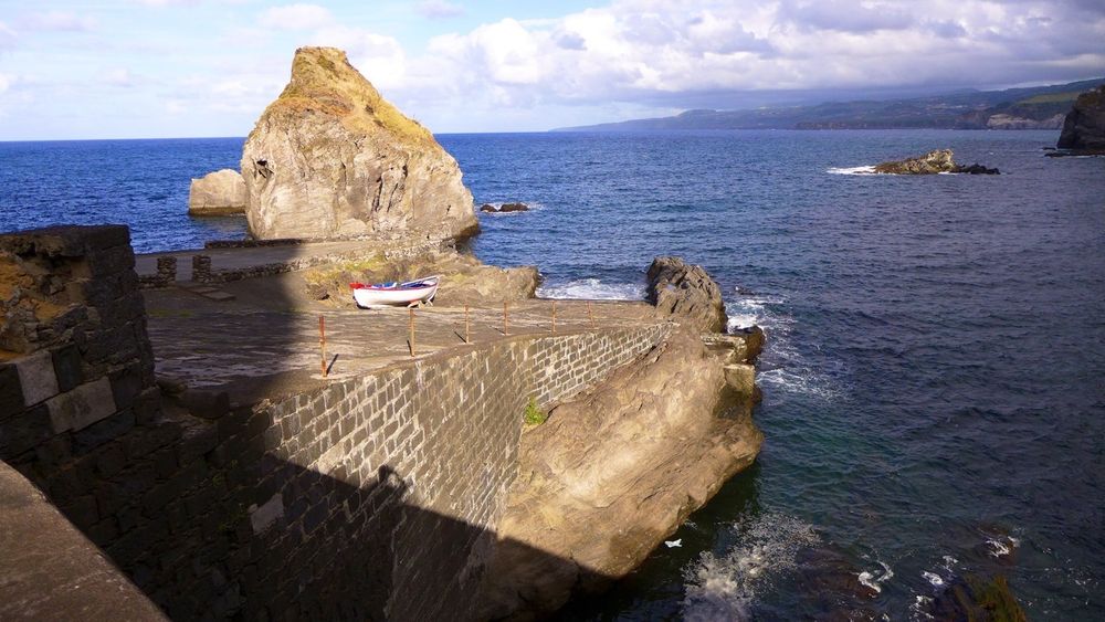 You can see an old, dilapidated ramp made of concrete bricks leading gently into the Atlantic Ocean. On the ramp is a small, open wooden fishing boat. Next to and behind the ramp, a large rock protrudes from the sea, protecting the entrance to the ramp from high waves. In the background on the right is the steep, rugged north coast of the island. As is often the case in the Azores, clouds have formed over the island, while the ocean is almost cloudless.